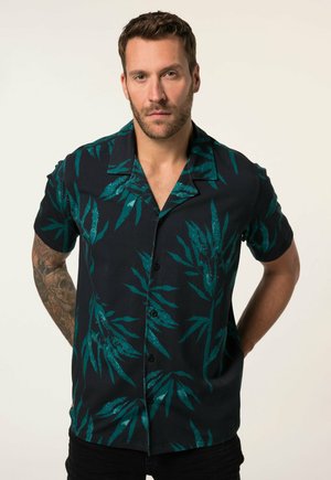 Uomo con capelli castani corti e barba, indossa una camicia nera a maniche corte con motivi di foglie verdi, in piedi con le mani dietro la schiena.