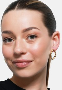 Gulløreringer i en organisk, uregelmessig form. Glatt metall med en polert finish, båret av en person med en nøytral bakgrunn.