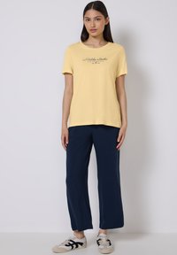 T-shirt en coton jaune à manches courtes, avec un logo en script noir. Associé à un pantalon large bleu marine et des baskets claires.