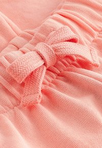 Szczegóły koralowo-różowego ubrania z miękkiej, teksturowanej tkaniny z marszczonymi miejscami i tkanym noskiem kokardy. Cały projekt jest gładki i elastyczny.