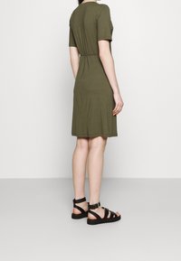 Robe olive à manches courtes avec une taille cintrée et une jupe évasée. Portée avec des sandales noires à lanières, sur un fond gris clair.