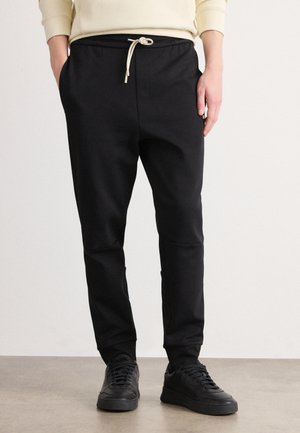 Person wearing black jogger pants with drawstring waist and black sneakers, standing with one hand in pocket against plain background.
