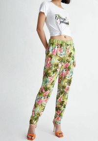 Woman wearing floral tropical print pants, a white knotted crop top with "Tropical Paradise" text, and orange high-heeled sandals.