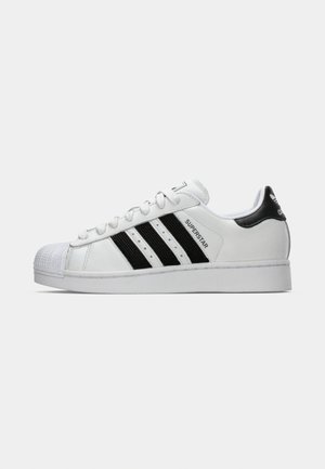 White sneaker with black stripes and accents, featuring a textured toe cap, round laces, and the "Superstar" logo on the side.