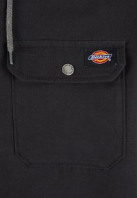 Svart bomullsstoff med en lomme, utstyrt med en rund metallknapp og en fargerik "Dickies" merkeetikett. Enkelt og funksjonelt design.