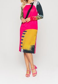 Vestido colorido hasta la rodilla con cuerpo rosa, mangas con patrones verdes y azules, detalles amarillos y líneas gráficas negras. Combinado con tacones rosas.