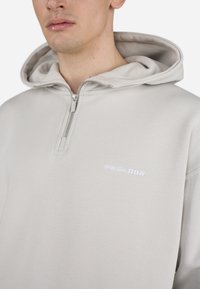 Svetlo siva pulover kapuca iz mehkega materiala ima pol-zip zapClosure, nastavljivo kapuco in belo vezeno logo "PEGADOR" na prsih.