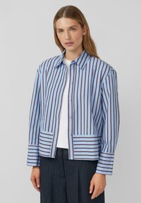 Camicia a righe blu e bianche con strisce marroni; presenta un colletto e una tasca frontale. Il tessuto sembra leggero e testurizzato.