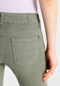 Pantalones de algodón verdes con un diseño ajustado, bolsillos traseros con un patrón de ola bordado en hilo claro y una pequeña etiqueta de logo.