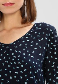Un haut marine avec un décolleté en V, orné d'un motif de formes circulaires bleu clair. Tissu doux à manches courtes, présentant une texture lisse.