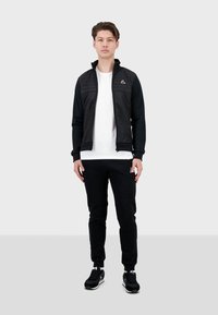 Giacca nera con zip e parte anteriore trapuntata, abbinata a una t-shirt bianca e pantaloni jogger neri. Scarpe sportive completano l'outfit.
