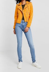 Suede oranžová motorkářská bunda s zipem a kapsami, v kombinaci s světle modrými skinny džíny a bílými teniskami.