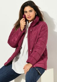 Burgunderrød puffjakke med quiltet design, høj krave og trykknaplukninger. Stylet med en hvid skjorte og mørke jeans.