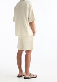 Ensemble en beige avec chemise à texture et short. Coupe ample, manches courtes et poches latérales sur le short. Porté avec des sandales sombres.