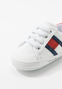 Zapatilla deportiva de cuero blanco con detalles en azul y rojo, punta redonda, cordones planos blancos y detalle de logo en el lateral. Textura lisa en toda la superficie.