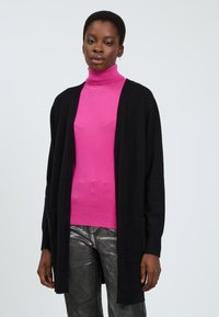 Zwarte lange cardigan met open voorkant, gedragen over een felroze coltrui en metallic, glanzende broek. Inclusief twee voorzakken.