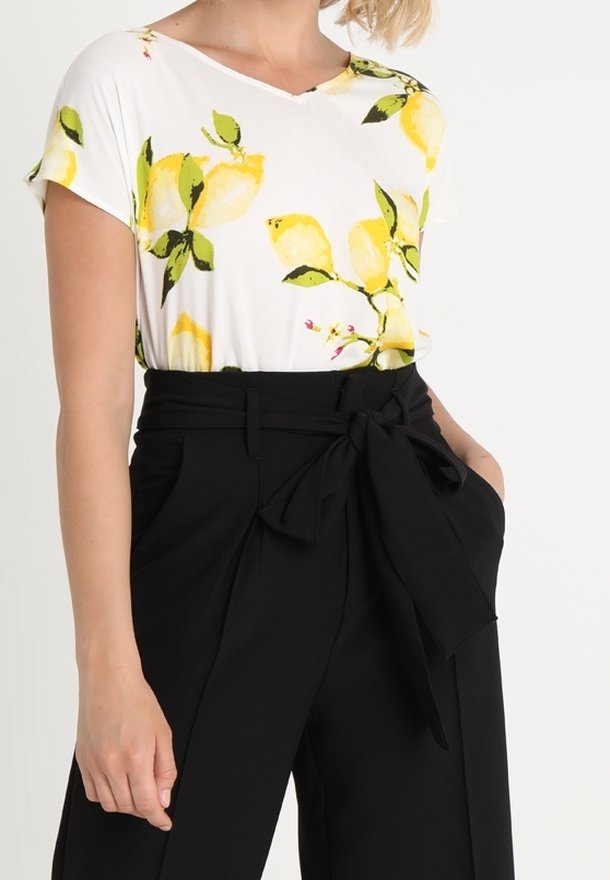 Femme portant un haut blanc à manches courtes avec un imprimé citron jaune et un pantalon noir taille haute avec une ceinture en tissu nouée.
