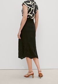 Jupe noire mi-longue avec un ourlet festonné, ornée de détails brodés. Portée avec un chemisier à motifs et des talons marron à brides.