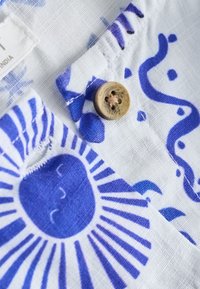 Lekka tkanina z niebieskim słońcem i abstrakcyjnymi wzorami, z jasnobrązowym guzikiem czterodziurkowym przyszytym beżową nicią.