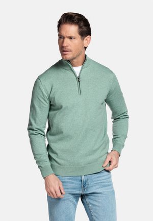 Man draagt een lichtgroene trui met ritssluiting en een witte blouse, gecombineerd met lichtblauwe spijkerbroek, en staat tegen een effen witte achtergrond.