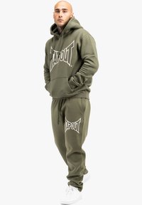 Olivengrønt hættegenser og sweatpants-sæt med et stort hvidt "TAPOUT" logo. Stoflooket ser blødt ud, med mansjetter og snoretræk detaljer. Bæres med hvide sneakers.