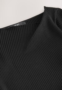 Gros plan d'un tissu en maille côtelée noire avec un col carré et une étiquette de marque partiellement visible à l'intérieur.