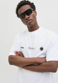 Hvid t-shirt med et sort solsymbol og tekstgrafik. Modellen bærer spejlblanke sorte solbriller og har kort, struktureret hår.