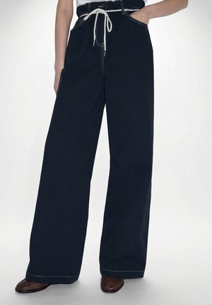 Person trägt hoch taillierte schwarze Hose mit weitem Bein und weißen Nähten, einen weißen Seilgürtel und braune geflochtene Schuhe, eine Hand in der Tasche.