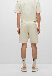 Muž v béžových šortkách, odpovídajícím tričku s nápisem "Dobrý vkus, žádný odpad", pruhovaných ponožkách a bílých teniskách s oranžovými detaily na patách, stojí.