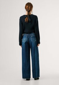 Mujer con el cabello trenzado atado con una cinta negra, vestida con un suéter de punto negro y jeans anchos de mezclilla, de pie contra un fondo simple.