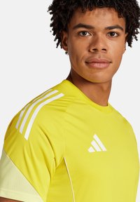 Maillot de sport jaune à manches courtes, avec un design à trois rayures blanches au niveau des épaules et un logo. Texture de tissu lisse et léger.