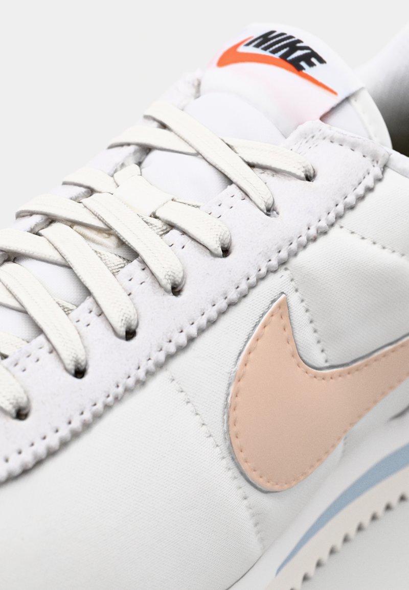 Baskets Nike blanches avec une tige en cuir lisse, un logo swoosh en pêche pastel, des coutures ton sur ton et des lacets plats classiques.