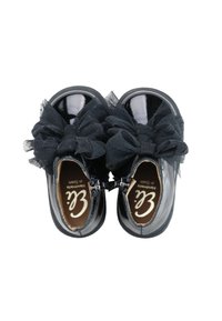 Zapatos de charol negro con dos grandes lazos de tul negro en la parte delantera, con cierre de cremallera y una plantilla marrón con marca.