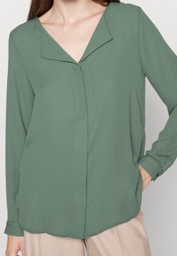 Blouse verte à manches longues avec un col en V, fabriquée en tissu lisse. Présente une fermeture à boutons centrée et un ourlet arrondi.