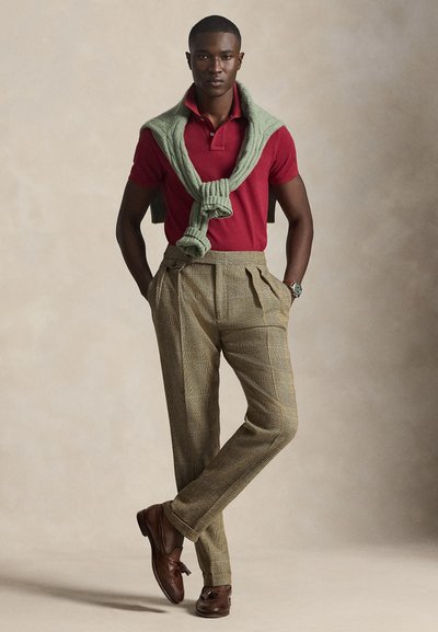 Άνδρας που φοράει κόκκινο πόλο, πράσινο πουλόβερ ριγμένο στους ώμους, καρό παντελόνι ψηλής μέσης, καφέ loafers με φούντες, στέκεται με τα χέρια στις τσέπες.