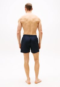 Μπλε σκούρο μαγιό με ελαστική μέση, πίσω τσέπη και λεπτομέρεια αποστράγγισης με οπή. Το μοντέλο εμφανίζεται από πίσω, ξυπόλυτο, σε λευκό φόντο.