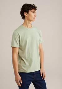 Šviesiai žalia medvilninė t-marškiniai su trumpomis rankovėmis, apvali kaklo išpjova, laisvas kirpimas ir minimalūs siūlės išsiskiria, derinami su tamsiai mėlynomis džinsinėmis kelnėmis.