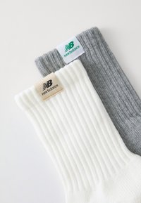 To sæt New Balance-sokker: et hvidt ribbet sæt med en beige etiket, og et gråt ribbet sæt med en grøn etiket, begge bløde og strukturerede.