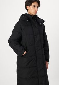 Svart vadderad lång kappa med huva, med en quiltad design, sidofickor, elastiska ärmslut och dragkedjestängning fram.
