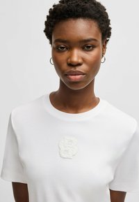 T-shirt bianco in cotone con un design logo testurizzato sulla parte superiore sinistra. Maniche corte e scollatura rotonda classica. Tessuto morbido.