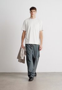 Λευκό βαμβακερό T-shirt με κοντά μανίκια, συνδυασμένο με χαλαρά γκρι τζιν. Το μοντέλο κρατάει ένα ριγέ γκρι πουκάμισο και φοράει μαύρα παπούτσια.