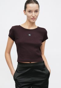 Camiseta corta color burdeos con cuello redondo y mangas cortas, con un parche de logo negro en el centro. Tejido suave y elástico de mezcla de algodón.