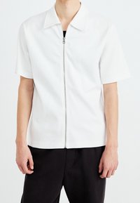 Chemise blanche à manches courtes en tissu lisse, dotée d'une fermeture éclair complète à l'avant et d'un col classique, associée à un pantalon noir.