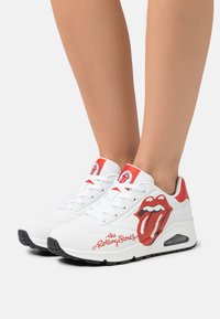 Vita sneakers med röda detaljer, med en framträdande Rolling Stones-logga i röd och svart på sidan. Materialet verkar vara syntetiskt med texturerade detaljer.