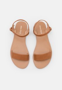 Un paio di sandali piatti color beige con cinturini alla caviglia e una sola fascia larga sulla punta, con il nome del marchio stampato sulle suole rivolte verso l'alto su uno sfondo bianco.