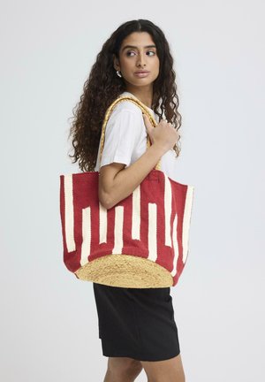 Donna con capelli lunghi e ricci che indossa una camicia bianca e una gonna nera, che porta una grande borsa tote intrecciata rossa e beige sulla spalla.