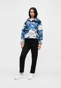 Γυναίκα που στέκεται, φοράει μπλε και λευκό φλις μπουφάν με σχέδια, μαύρο παντελόνι, λευκές κάλτσες και λευκά αθλητικά παπούτσια, σε απλό φόντο.