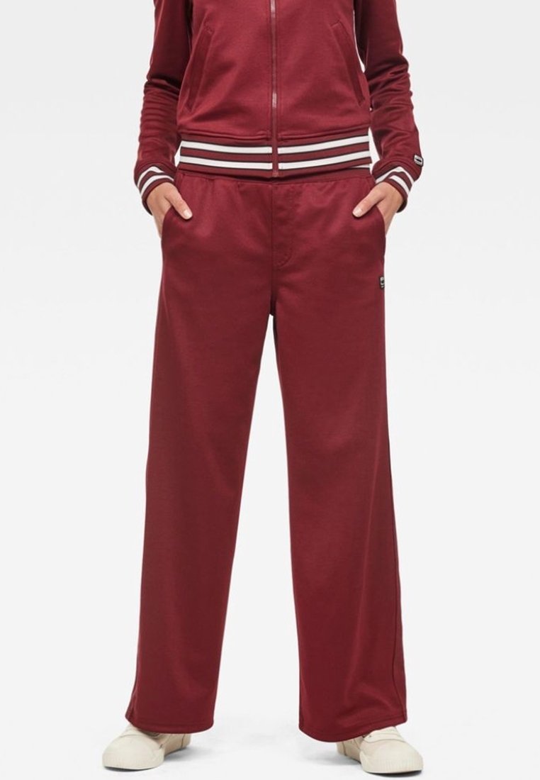 Personne portant un pantalon large bordeaux et une veste zippée assortie avec des poignets et une ceinture rayés blancs, mains dans les poches, debout sur des chaussures blanches.
