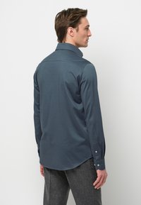 Maglietta a maniche lunghe di colore blu scuro con tessuto morbido, orlo arrotondato e polsini con bottoni. Presenta un colletto classico e una texture liscia.
