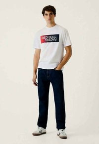 T-shirt branca de algodão com gráfico "RED BULL RACING" em vermelho e azul, combinada com jeans azul-escuro e ténis brancos.